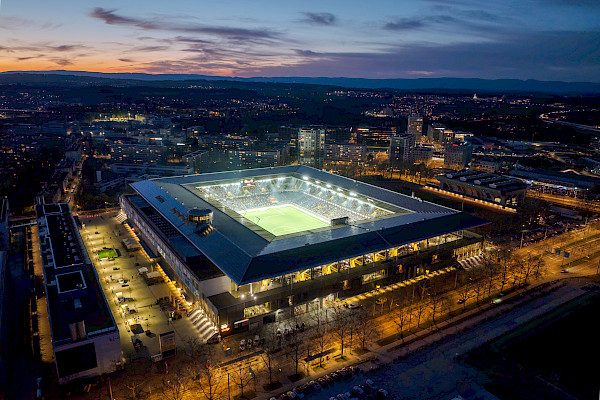 Angled drone view of the brightly lit Wankdorf Stadium at night, surrounded by the city of Bern with hills on the horizon.