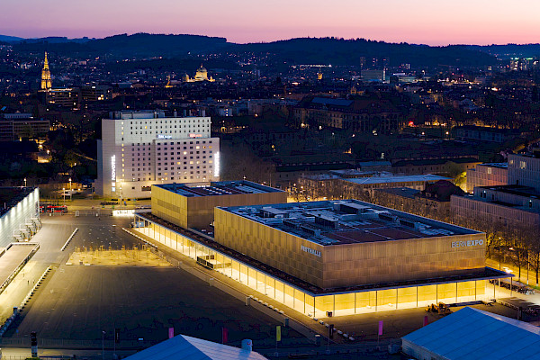 Aerial view of the BERNEXPO area and the Festhalle in Bern at dusk, showing hotels, exhibition halls, and the city skyline in the background.