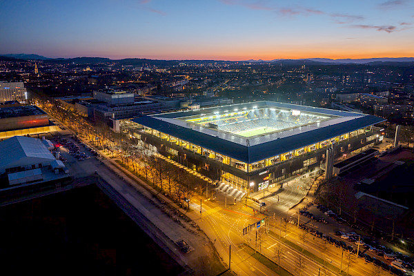 Aerial view of the illuminated Wankdorf Stadium in Bern at dusk, surrounded by city lights and traffic.